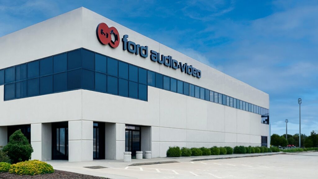 Exterior of the Ford Audio-Video office building with modern architecture, tinted windows, and landscaped grounds under a blue sky.