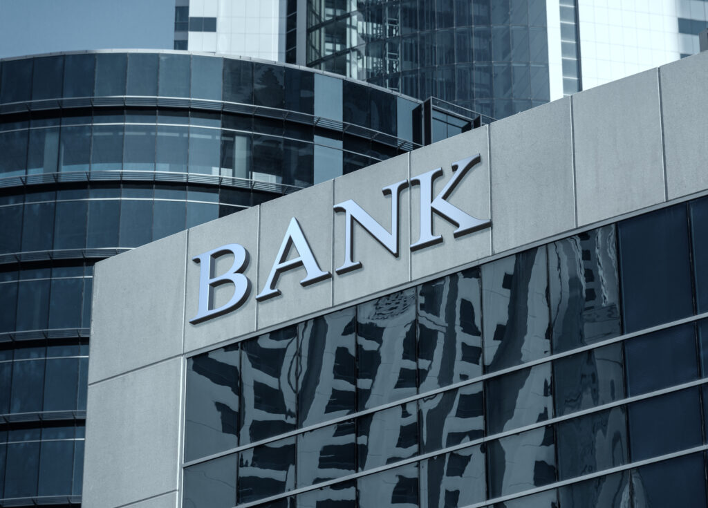 Close-up of a modern bank building with a prominent BANK sign and reflective glass facade in an urban financial district.