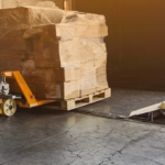 A worker steers a yellow manual pallet jack loaded with plastic-wrapped boxes up a metal ramp into a delivery truck at an industrial loading dock.