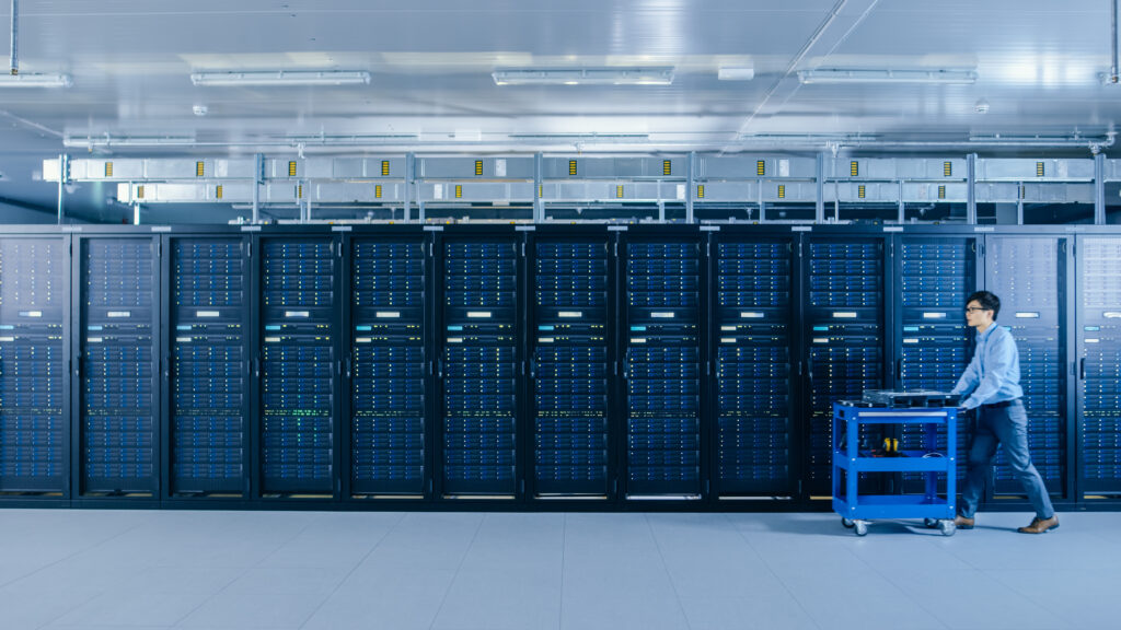 IT technician pushing a cart with server hardware through a modern data center aisle lined with active server racks.