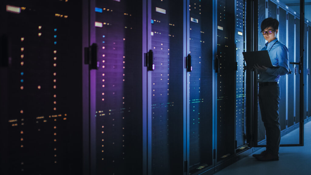 IT professional using a laptop beside server racks in a modern data center with blue status lights.