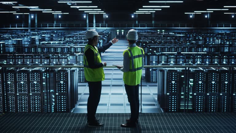 Two data center professionals in safety vests and hard hats stand on an elevated platform overlooking rows of server racks, representing infrastructure oversight, planning, and facility management.