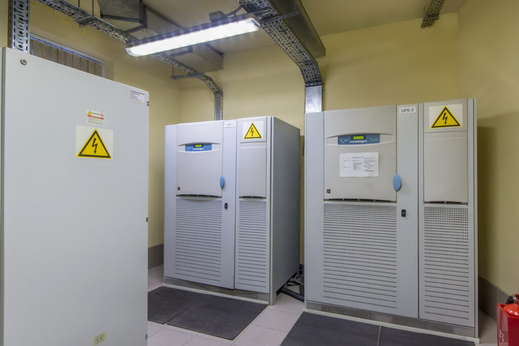 Electrical room with industrial UPS units, overhead cable trays, high-voltage warning labels, and safety mats in front of backup power equipment.
