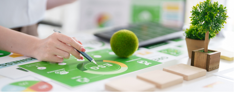 Hand pointing to an ESG reporting document on a desk with plants, wooden blocks, and office materials, representing sustainability metrics and regulatory compliance.