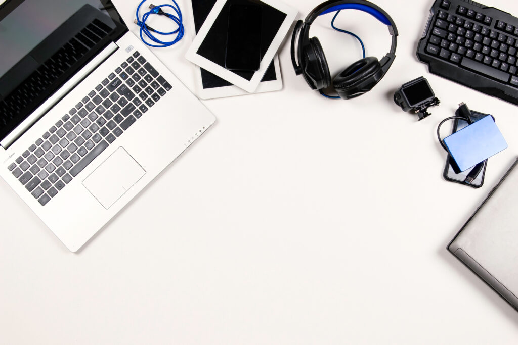 Arrangement of laptops, tablets, headphones, a keyboard, cables, and other tech accessories on a white background with open copy space.