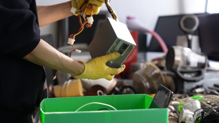 Person wearing yellow gloves sorting a computer power supply with bundled cables above a green bin in a workspace.