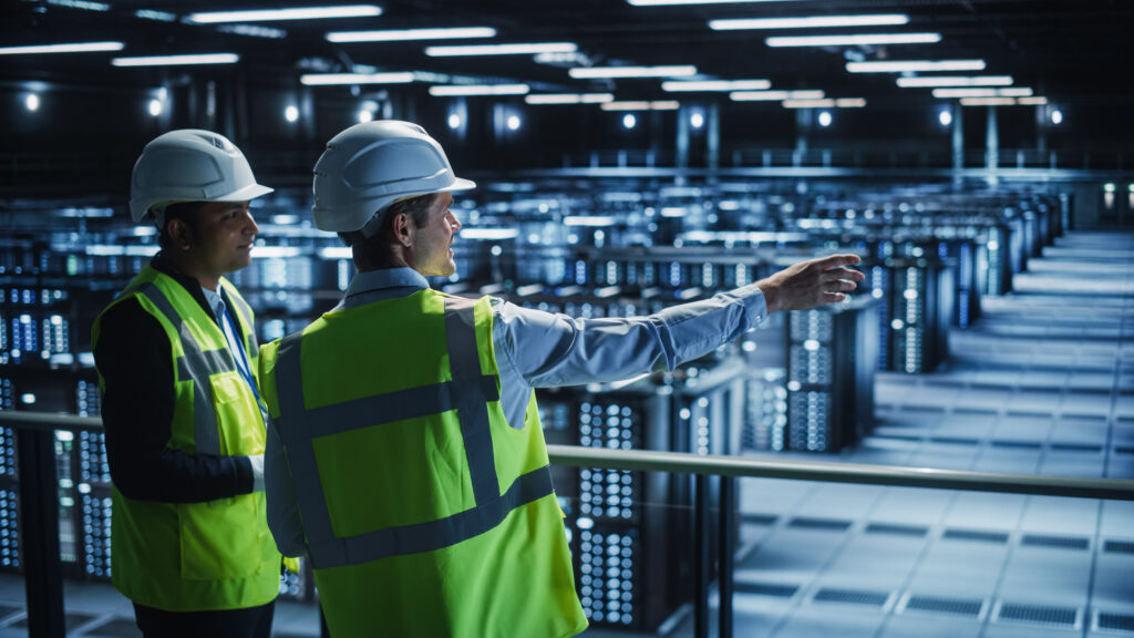 Two data center engineers in hard hats and safety vests overlooking server racks from an elevated walkway.