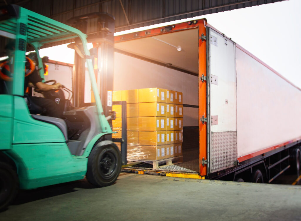 Forklift operator loading a pallet of wrapped cardboard boxes into a shipping truck at an industrial loading dock.