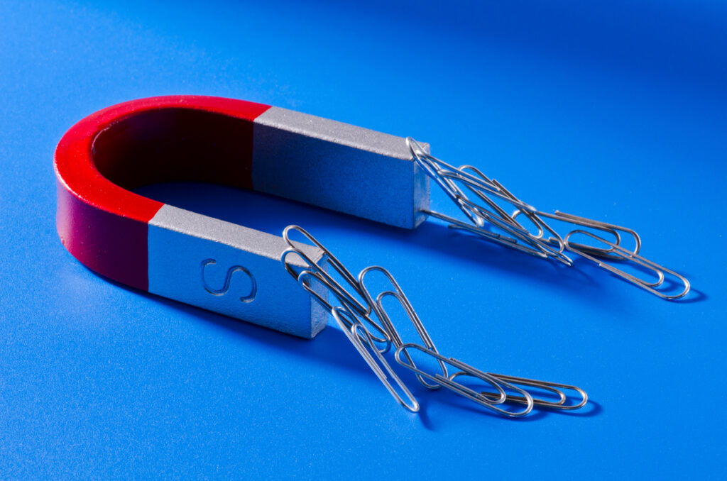 Red and silver horseshoe magnet on a blue surface attracting several silver paperclips to both poles, with some clips hanging in short chains.