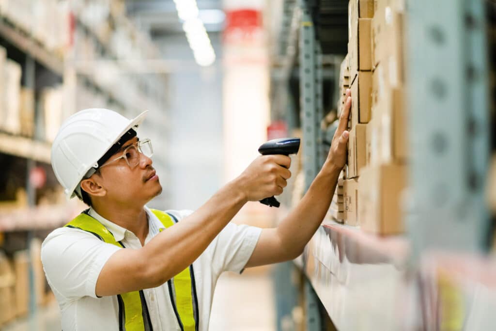 Warehouse worker in safety gear scanning stacked cardboard boxes with a barcode scanner on a storage shelf.