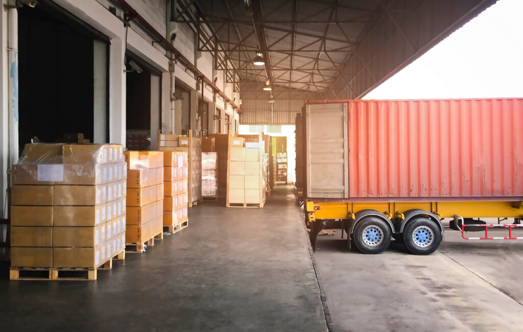 Warehouse loading dock with palletized cardboard boxes beside a shipping container truck.
