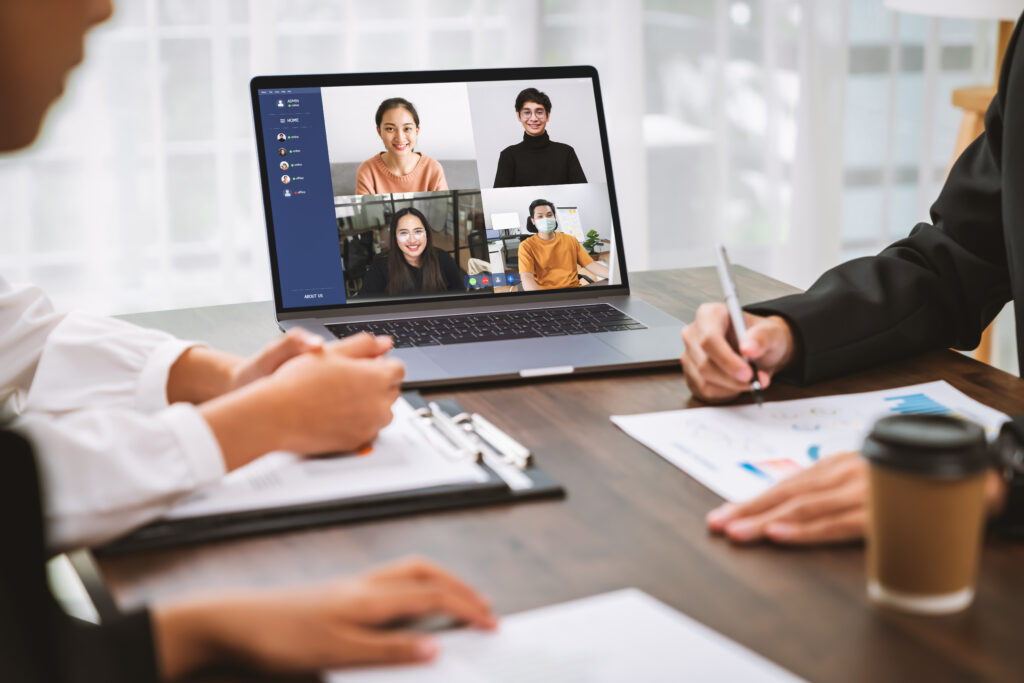 Two professionals review printed charts at a conference table while joining a laptop video call with four remote colleagues in a bright office, illustrating a hybrid business meeting.