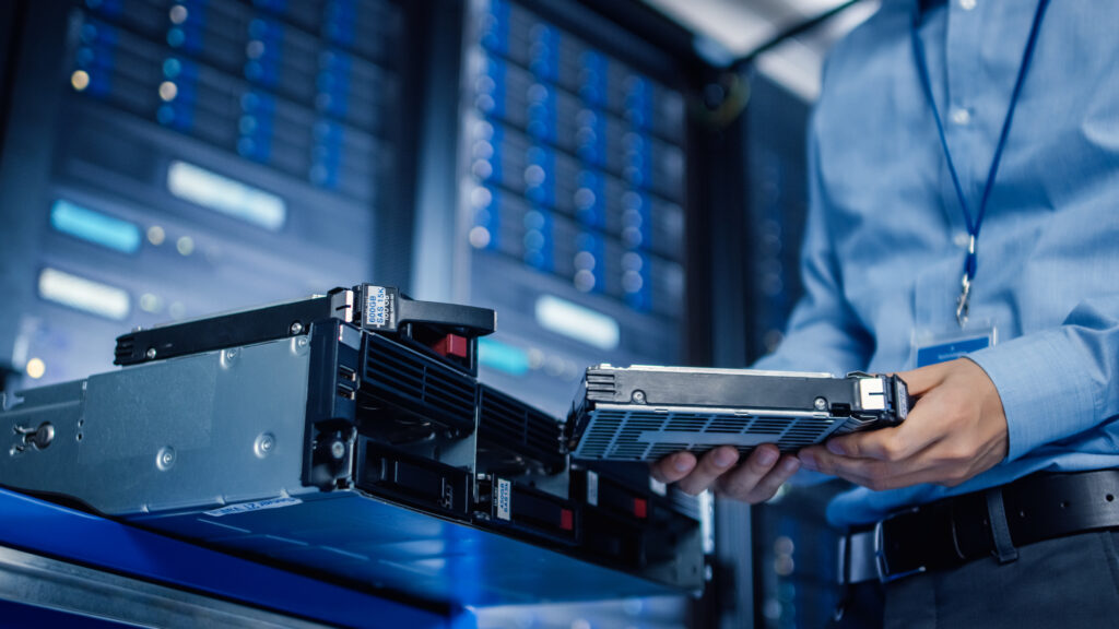 An IT professional in a blue shirt holding a 600GB SAS15K hard drive, with a server rack in the background, highlighting data center maintenance.