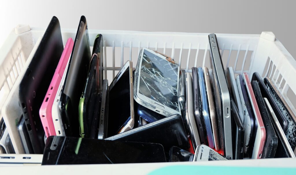 A white plastic crate filled with old, damaged smartphones and tablets, many with cracked screens and visible wear, symbolizing electronic waste ready for recycling.