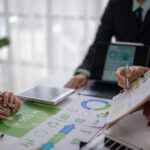 A business meeting scene with hands pointing at ESG (Environmental, Social, Governance) documents and charts on a white desk, emphasizing sustainability and corporate responsibility while managing e-waste for a remote workforce.