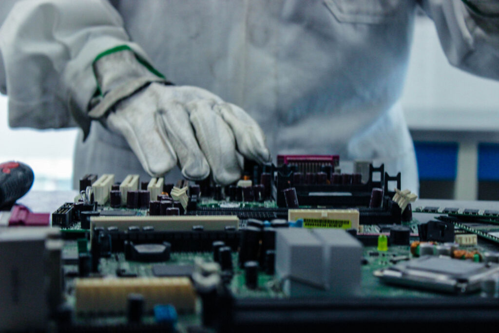 A close-up of a person in a lab coat and gloves working on circuit boards, focusing on intricate electronic components in a professional lab or workshop setting.