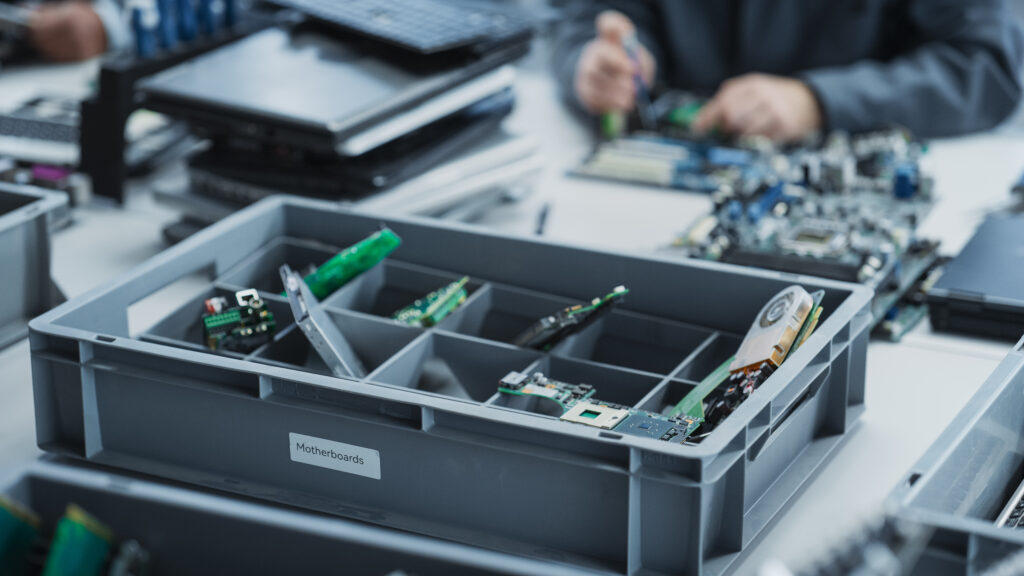 A gray plastic bin labeled "Motherboards" filled with green circuit boards, set on a workbench in an electronics recycling or repair workshop.