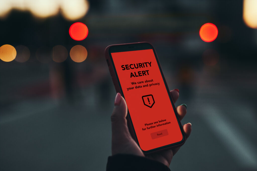 A smartphone displaying a red "SECURITY ALERT" screen about data and privacy, held by a person with blurred city lights in the background.